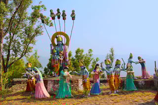 Large statue featuring Lord Krishna and Radha surrounded by gopis engaged in the traditional Raas Leela dance. The figures are positioned on a raised platform outdoors, with lush greenery and trees in the background, creating a joyful and serene atmosphere