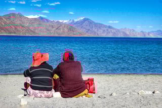 Two tourists sit beside the calm waters of Lake Pangong in Kashmir during daylight hours, gazing at the expansive lake and distant mountains. The scene is peaceful and serene, with clear blue skies and rugged natural surroundings creating a tranquil atmosphere
