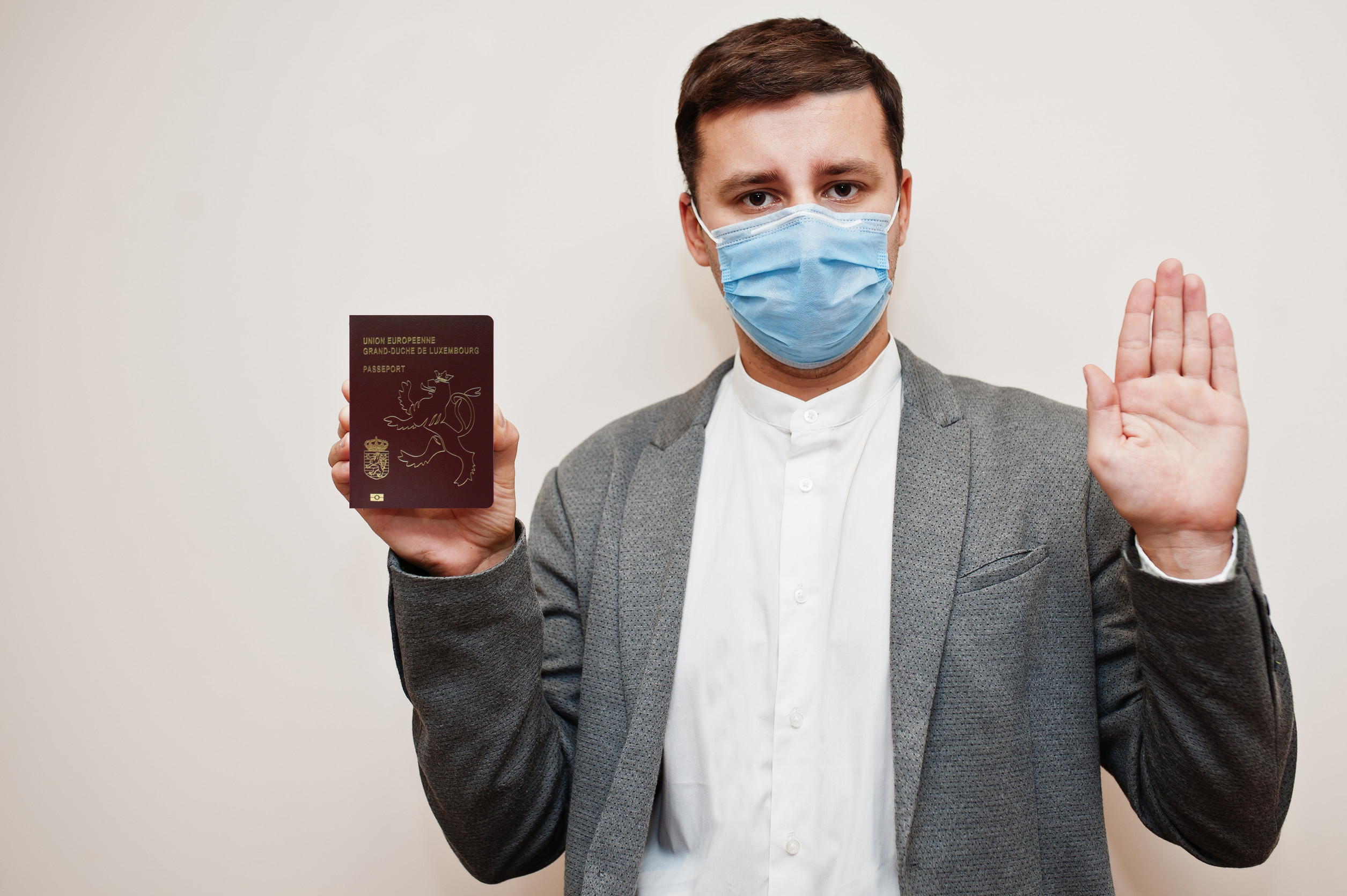 Europäischer Mann in formeller Kleidung und Gesichtsmaske zeigt luxemburgischen Reisepass mit Stoppzeichen-Hand