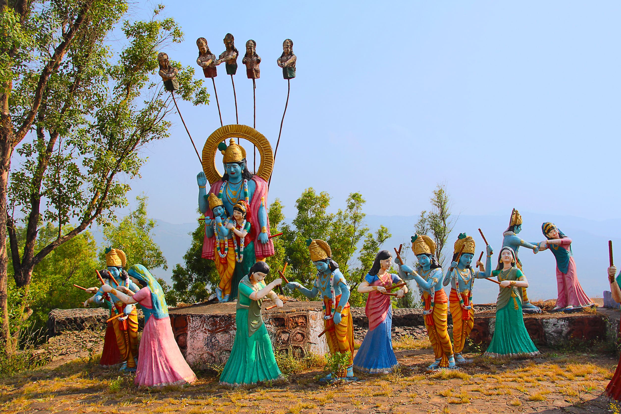 Grande statua che raffigura il Signore Krishna e Radha circondati dalle gopi impegnate nella tradizionale danza Raas Leela. Le figure sono all'aperto su una piattaforma rialzata con vegetazione e alberi sullo sfondo, creando un'atmosfera serena e gioiosa