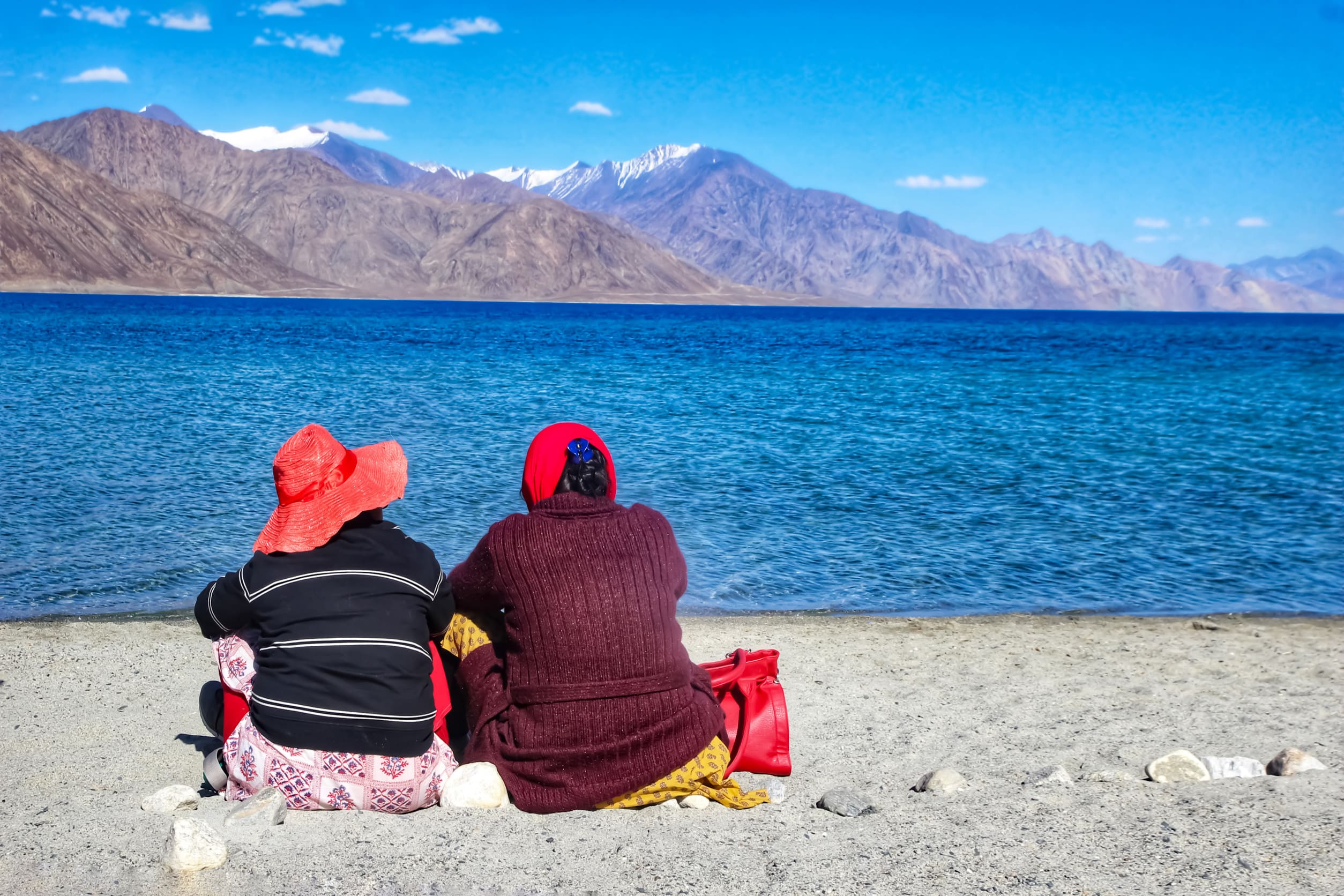 Zwei Touristen sitzen tagsüber am ruhigen Ufer des Pangong-Sees in Kaschmir und blicken auf das weite Wasser und die fernen Berge. Die Szene ist friedlich und ruhig, mit klarem blauen Himmel und einer rauen natürlichen Umgebung, die eine entspannte Atmosphäre schafft.